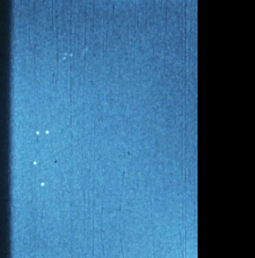 A frame of grainy blue sky from a 1952 16mm color film, with a loose cluster of four or five small bright points near the left of the frame.
