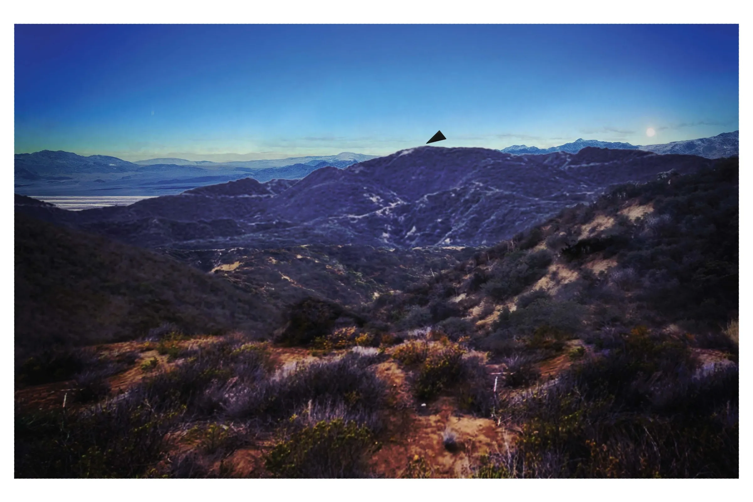 A landscape photograph of brush-covered hills at dusk, with distant ridges and a valley floor, and a small black triangle drawn above the central ridge line.